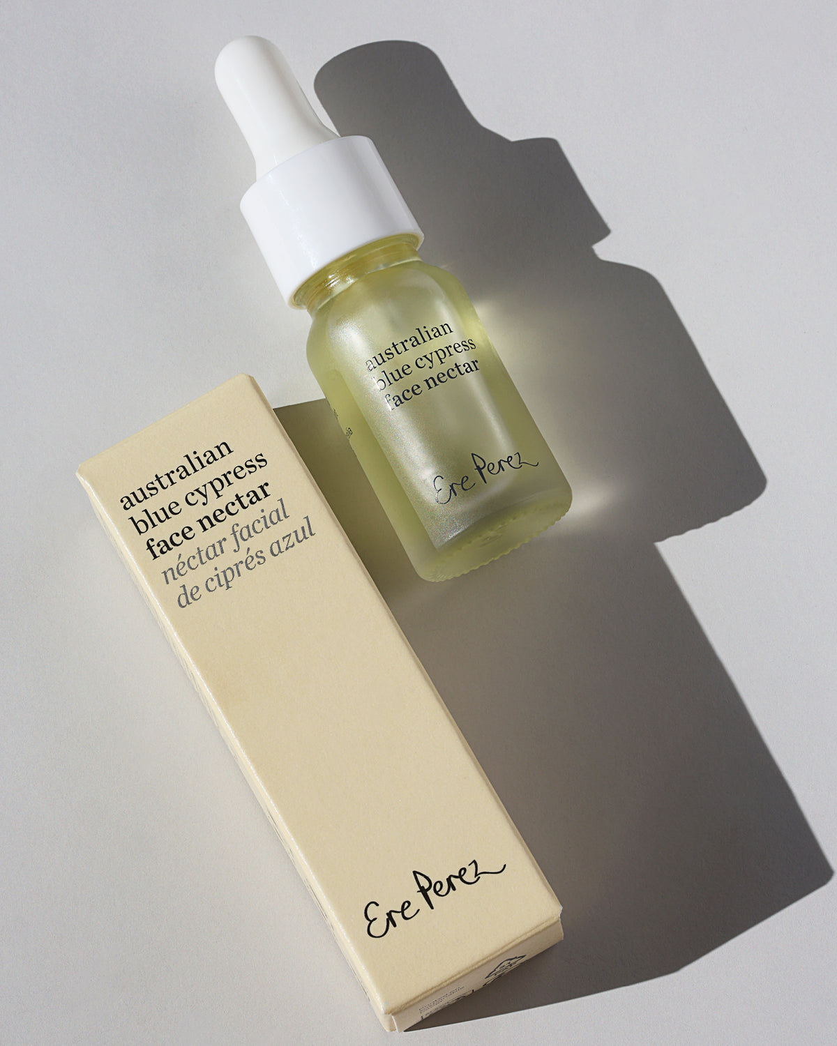 A bottle of Etc. Perez Australian Blue Cypress Face Nectar with a white dropper cap next to its cream-colored cardboard box, set on a light gray surface with a strong diagonal shadow