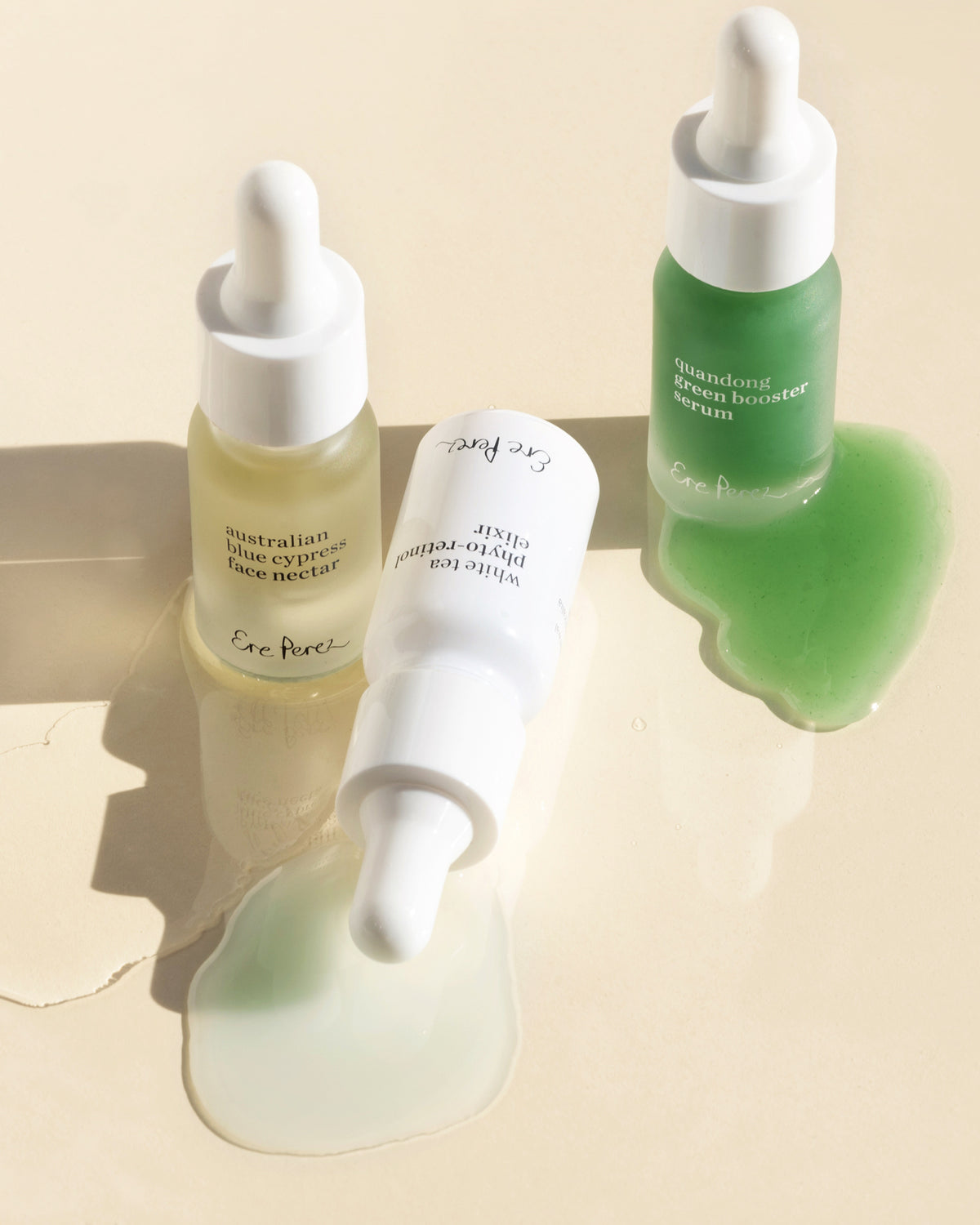 Three skincare dropper bottles from Ece Pivaci arranged on a warm beige reflective surface with liquid gels visible in the bottles and puddles. One bottle contains a pale yellow gel, one is white, and one is translucent green