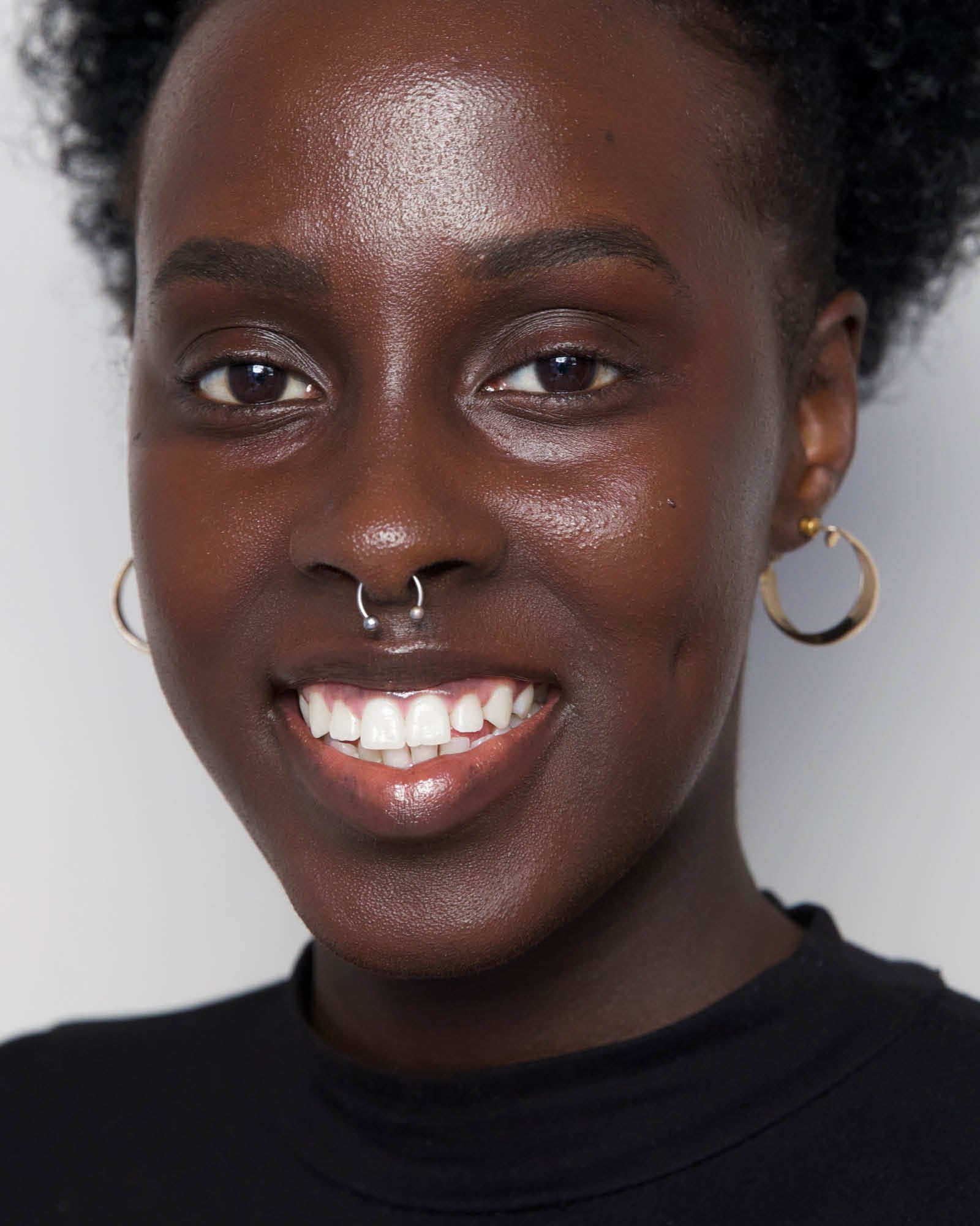 Close-up of a person with a nose ring and hoop earrings against a plain background