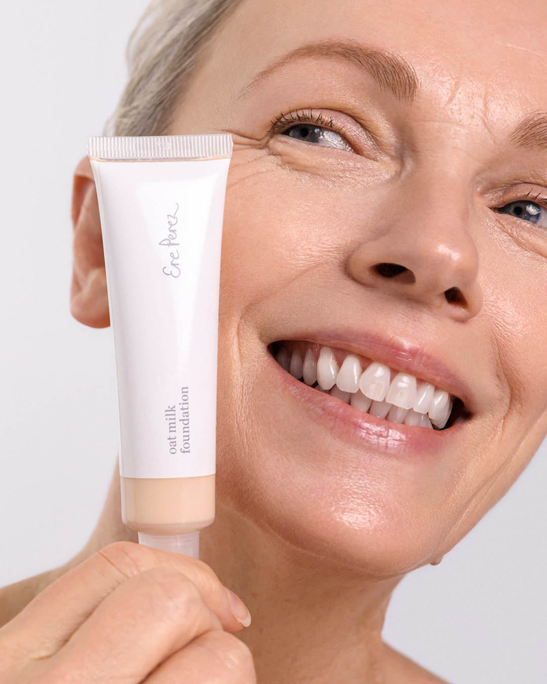 Woman holding a tube of foundation near her face against a light background,