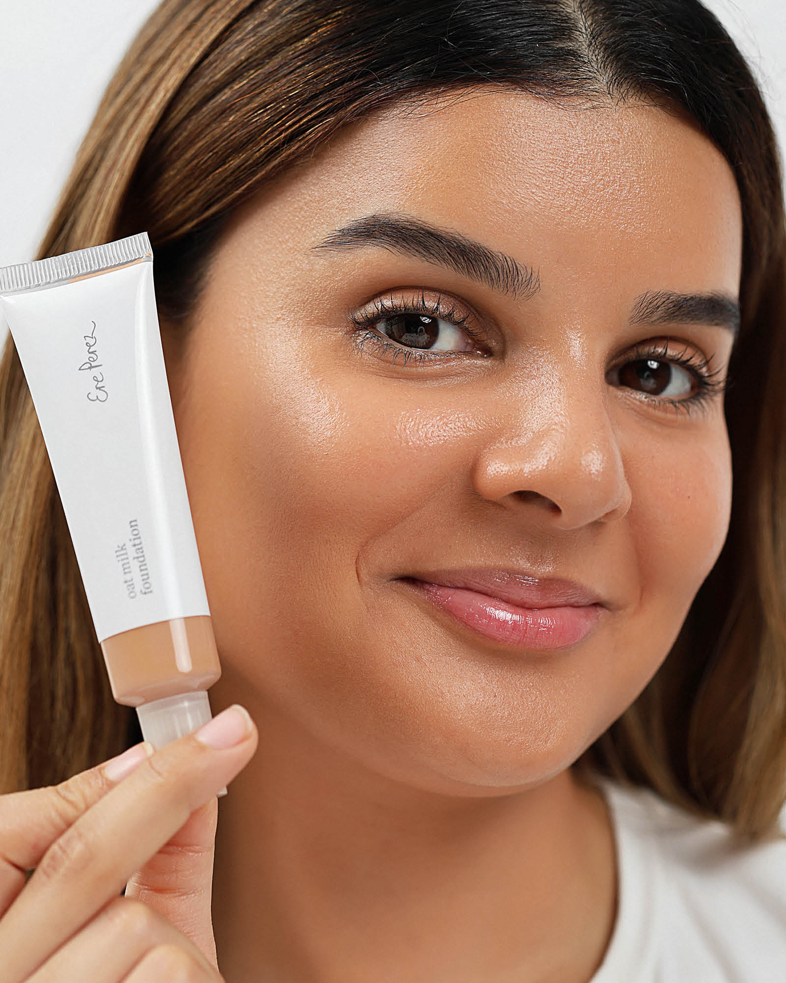 Woman holding a tube of Ere Perez foundation in shade Syrup against her face,