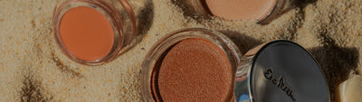 Three open pots of Ere Perez cream blush and bronzer arranged in the sand, displaying different shades.