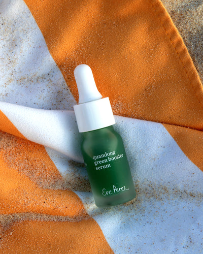A small dropper bottle of ere perez Green Booster Serum in a frosted dark green glass with a white dropper cap, styled on a white and orange striped beach towel with sand scattered around it 