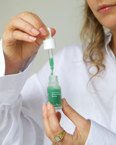 A close-up of a person holding a small glass dropper bottle of green serum, with the dropper lifted to release the green liquid. The person is wearing a white blouse and gold rings, against a soft light gray background 