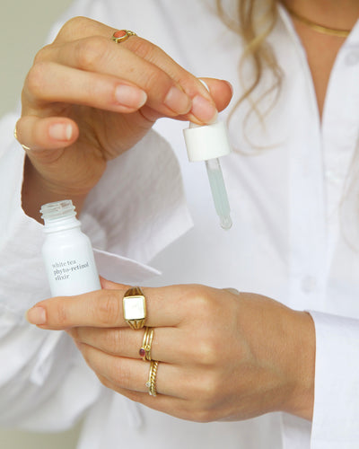 A close-up of a person in a white shirt holding a small white bottle and lifting the dropper of a clear liquid, with gold rings visible on both hands 